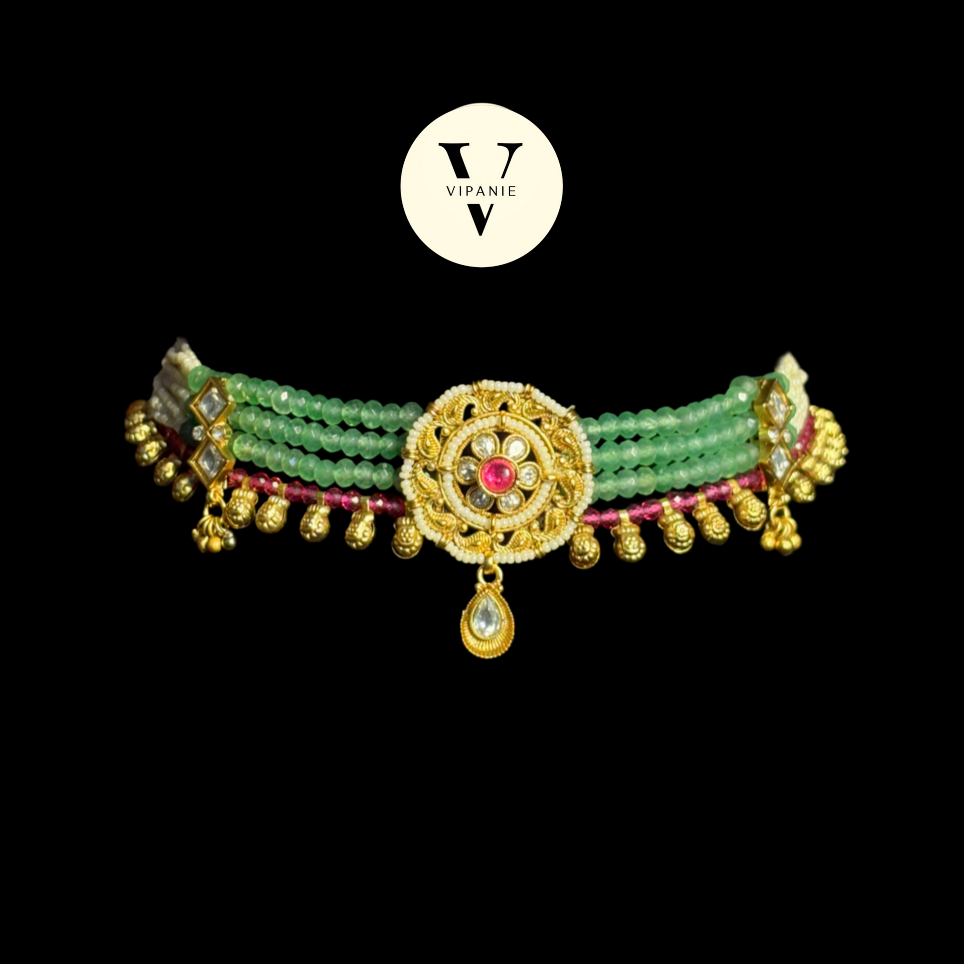 A traditional Indian choker necklace by Vipanie, featuring green beads, gold-plated metal elements, a central gold medallion with a red stone, and a teardrop pendant, set against a solid black background.