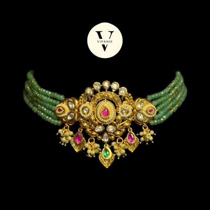 An ornate gold-plated choker necklace with green beaded strands, white faux pearls, and pink and green gemstone accents, displayed on a solid black background with a white Vipanie logo above.