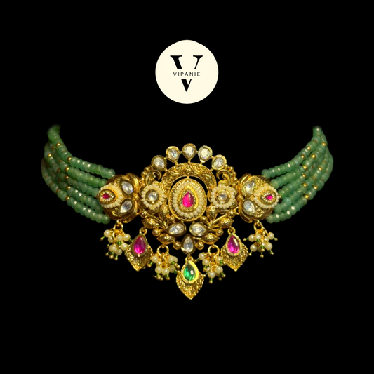 An ornate gold-plated choker necklace with green beaded strands, white faux pearls, and pink and green gemstone accents, displayed on a solid black background with a white Vipanie logo above.