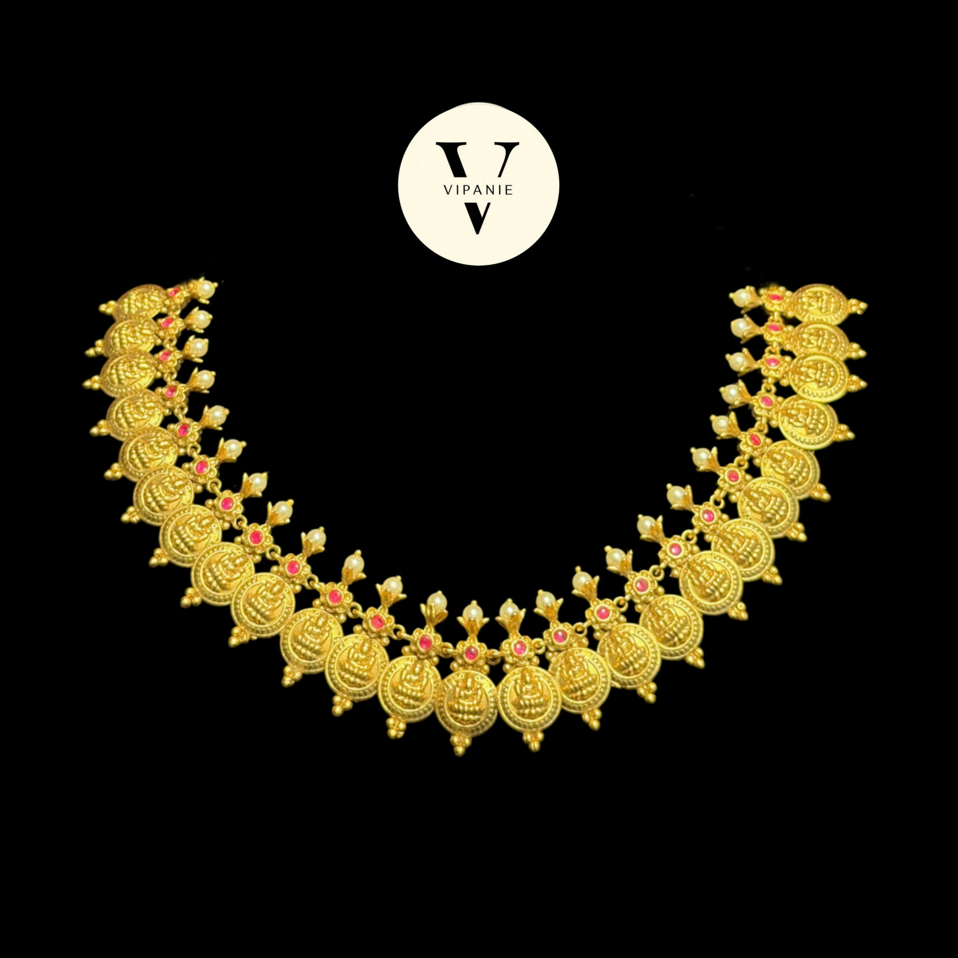 A gold-toned traditional Indian necklace with intricate filigree patterns and small pink stone accents, displayed on a black background with a circular Vipanie logo above it.